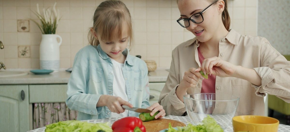Adorable little girl is helping mother in kitchen cutting vegetables for salad and talking making dinner together. Healthy nutrition, lifestyle and people concept.