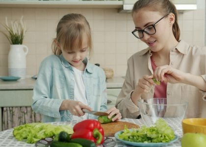 Adorable little girl is helping mother in kitchen cutting vegetables for salad and talking making dinner together. Healthy nutrition, lifestyle and people concept.