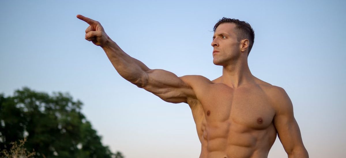 Shirtless muscular man posing and pointing outdoors with a clear blue sky background.